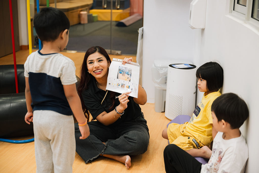 Awwa staf sitting down in a classroom set up in a small circle with 1 boy standing showing a picture, with 2 kids sitting down
